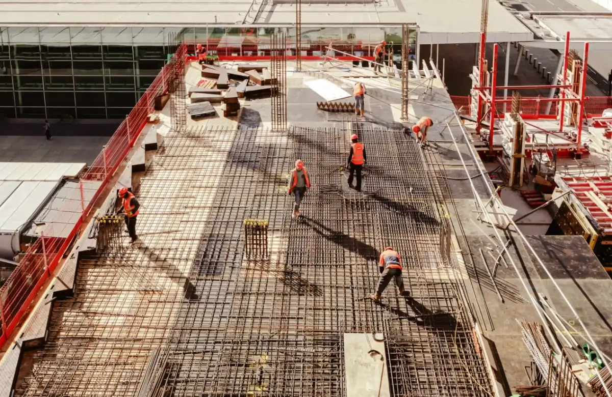 Construction workers in orange vests are working on a building's concrete framework. The site is elevated, with visible steel reinforcements and surrounding safety barriers.