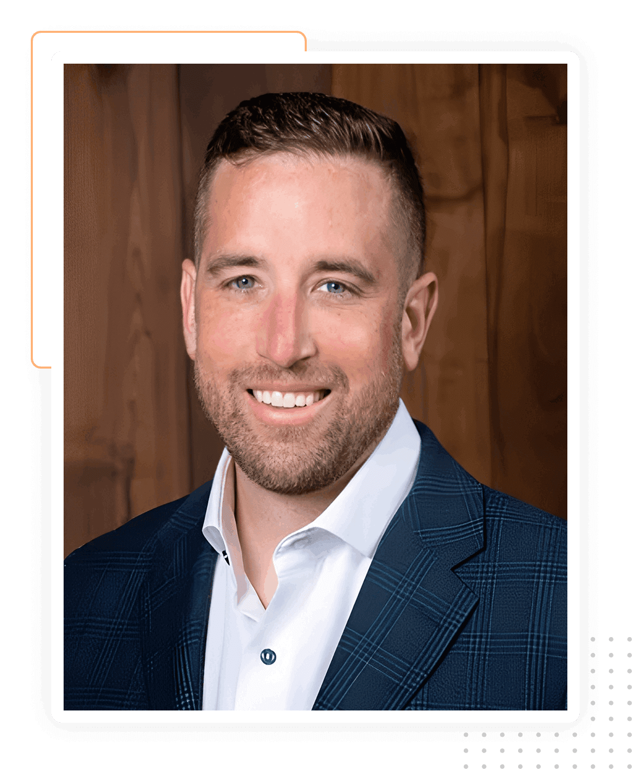 A man with short hair and a beard, wearing a dark checked blazer and white shirt, is smiling in front of a wooden background.