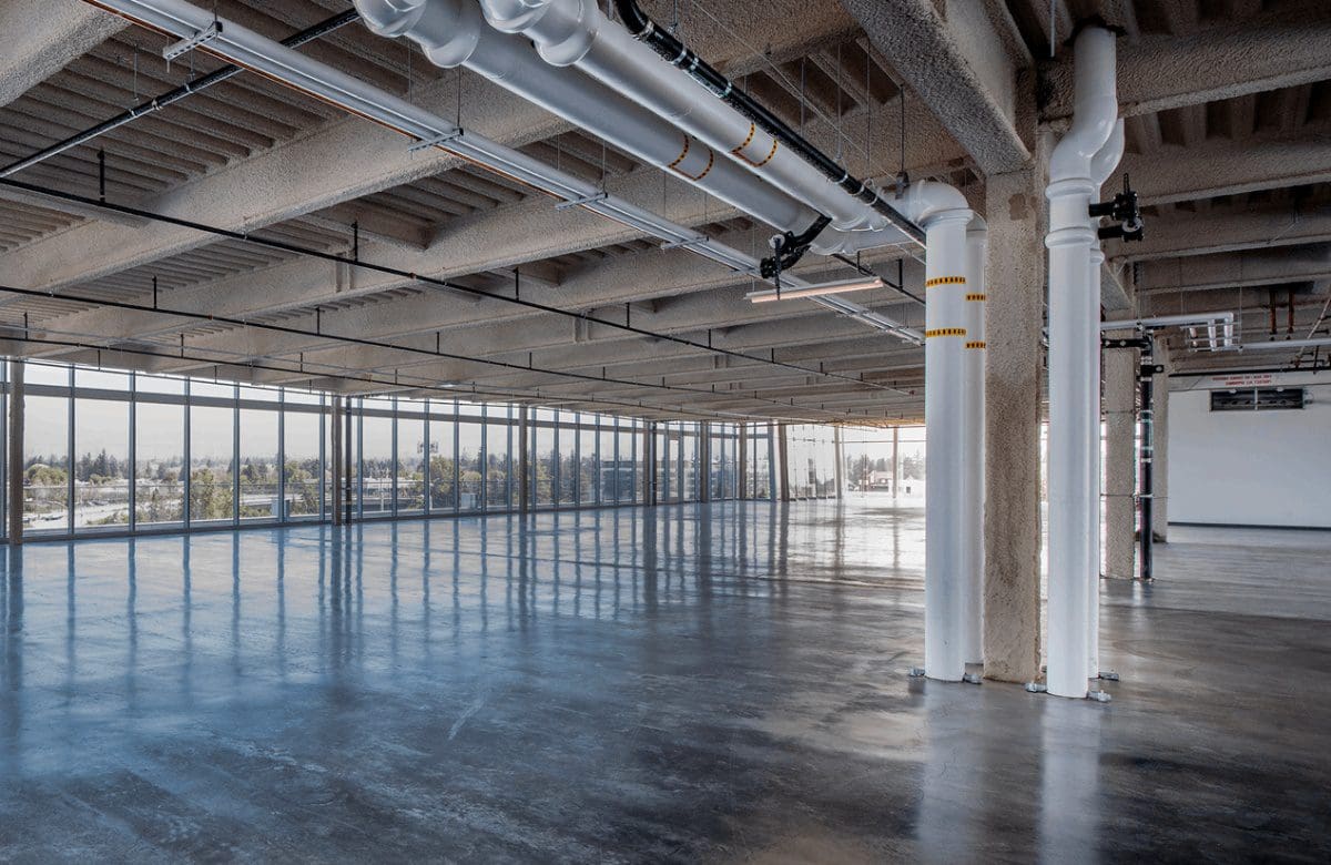 Empty industrial space with exposed ceiling beams, large windows, and polished concrete floor, featuring white structural pipes and a view of outdoor greenery.
