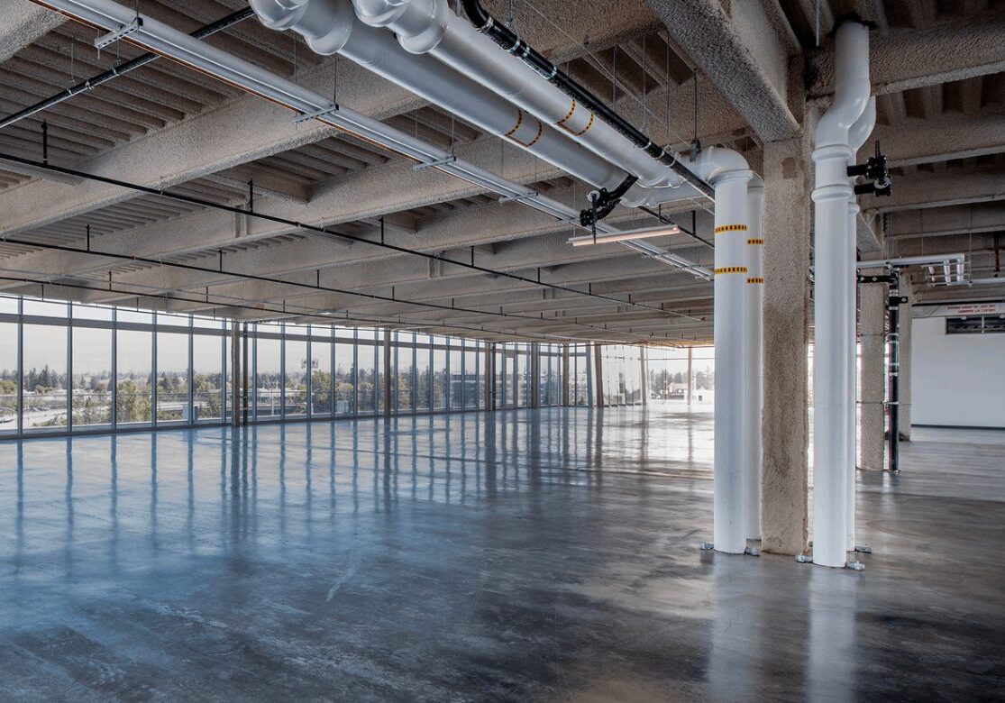 Empty industrial space with exposed ceiling beams, large windows, and polished concrete floor, featuring white structural pipes and a view of outdoor greenery.