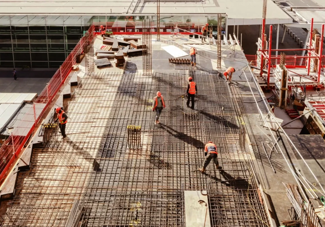 Construction workers in orange vests are working on a building's concrete framework. The site is elevated, with visible steel reinforcements and surrounding safety barriers.